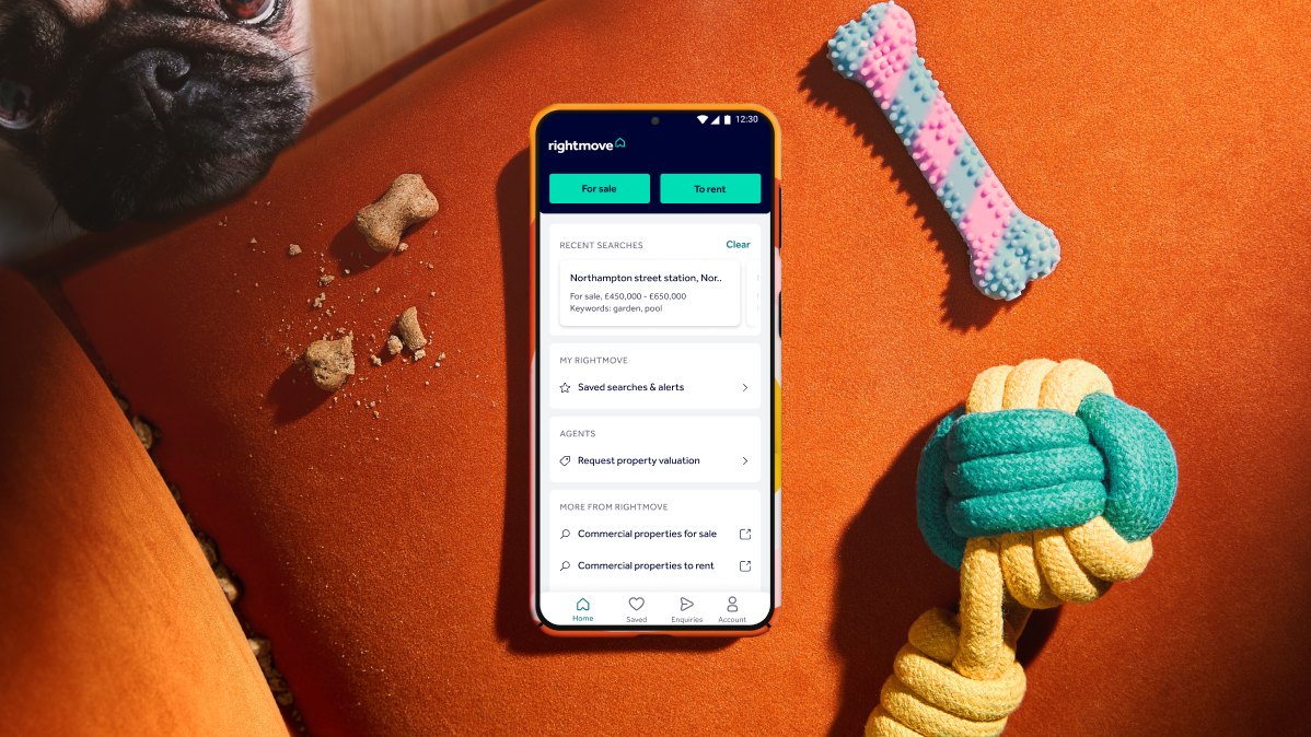 The Rightmove app's home screen on a phone lying on an orange sofa, with dog toys beside it and a pug looking in from the corner.